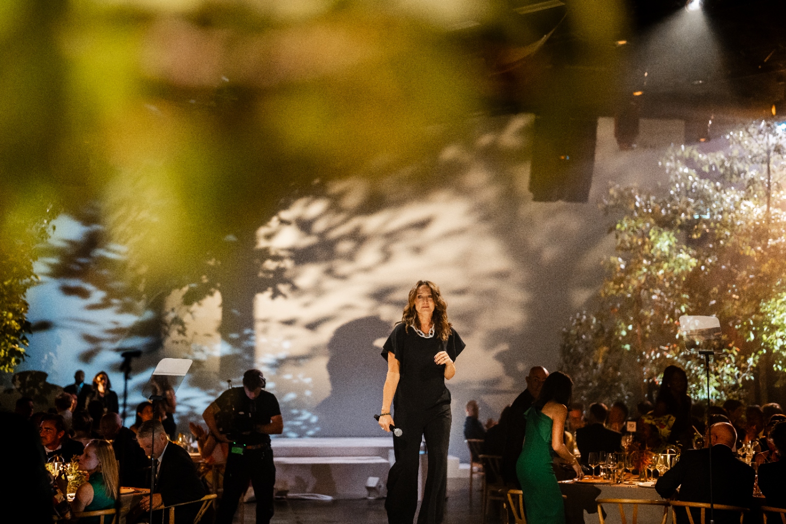 A speaker addressing guests at THE NAT GALA, framed through greenery with seated tables in the background
