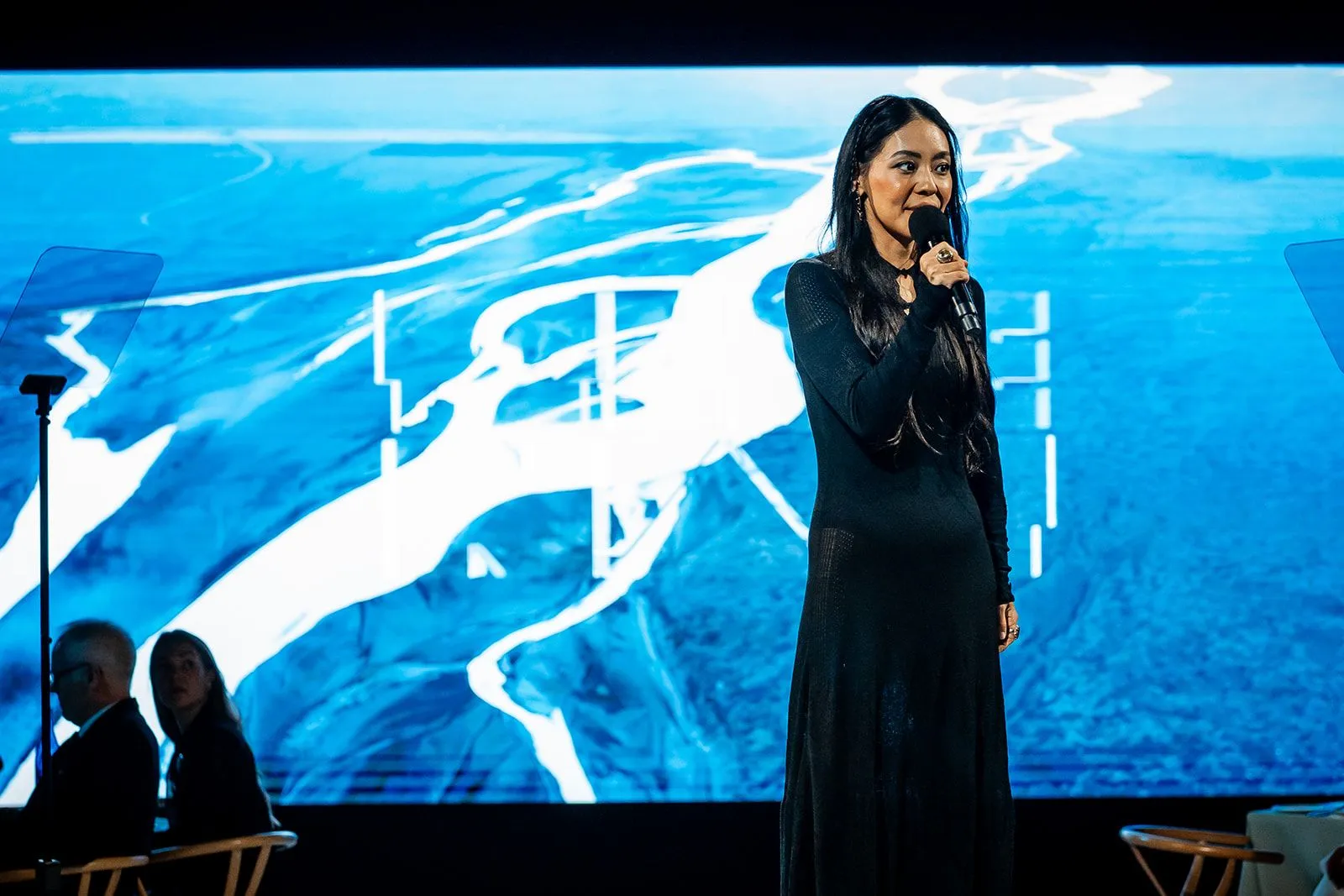 A speaker on stage in front of a projected aerial river delta.